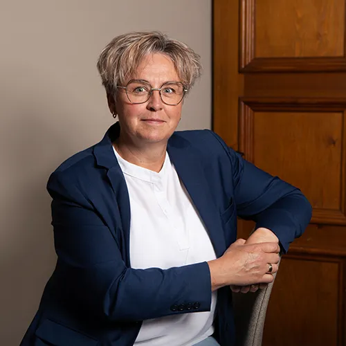 En person med glasögon i marinblå kavaj och vit skjorta sitter vid en trädörr och tittar in i kameran och utstrålar en lugn professionalism som påminner om någon man kan träffa på en begravningsbyrå Värnamo.