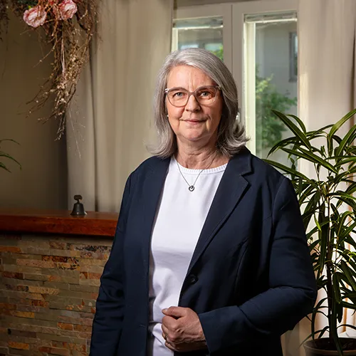 En äldre kvinna med grått hår och glasögon står inomhus, iklädd mörk jacka och vit skjorta, nära en växt. Hennes sammansatta hållning tyder på expertis i frågor som juridik, kanske kopplat till den lokala begravningsbyrån i Gnosjö eller Gislaved.