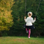 Ett barn som svänger på en gunga i en skogsmiljö. Hon har på sig en vit tröja, en svart kjol och rosa strumpbyxor. I bakgrunden syns gröna träd och en mjukt belyst gräsyta.