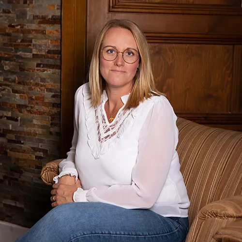 En kvinna med glasögon sitter i en soffa, iklädd vit blus och jeans, mot en träpanel och stenväggsbakgrund, som påminner om lugna utrymmen i Värnamos välrenommerade begravningsbyrå.