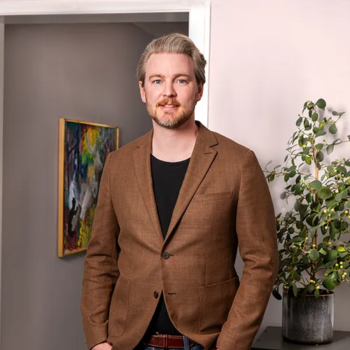 En man i brun kavaj står vid en växt, med en färgstark målning som påminner om den konstnärliga charm som ofta finns i Värnamos begravningsbyråer, vilket förstärker den fridfulla atmosfären.